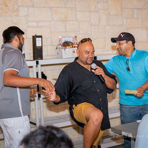 Three men are interacting indoors; one is holding a microphone while others assist him. There are shelves and a brick wall in the background.