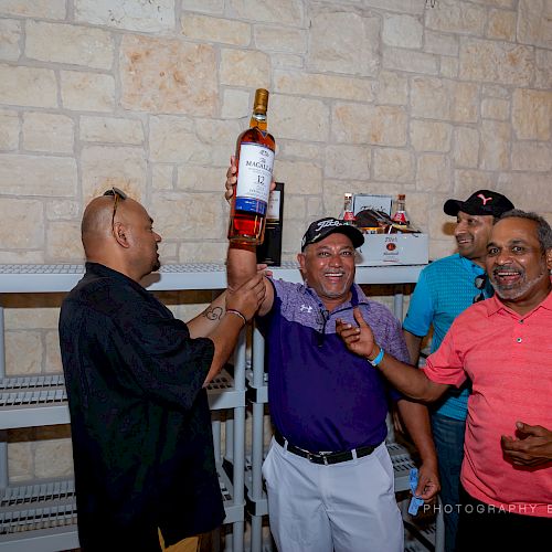 Four people are in a room with stone walls, laughing and holding a large bottle of alcohol. Two shelves are in the background.