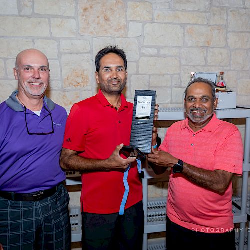 Three men are posing indoors, with one holding a trophy between them, all smiling at the camera.