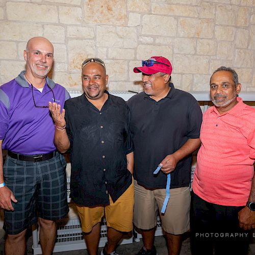 Four men are standing and smiling in front of a stone wall, with one making a hand gesture while others are dressed casually.