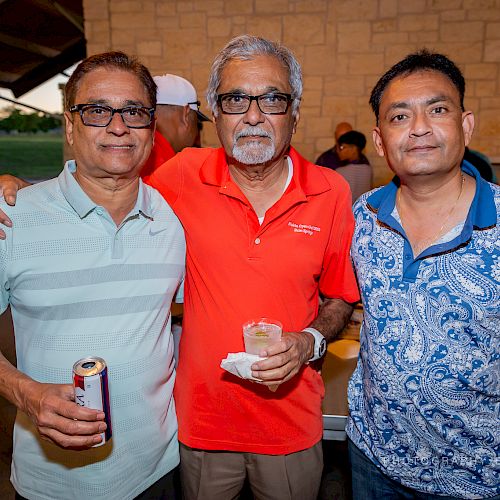 Three men posing together, one holding a can and another holding a drink, in a casual outdoor setting with smiles.