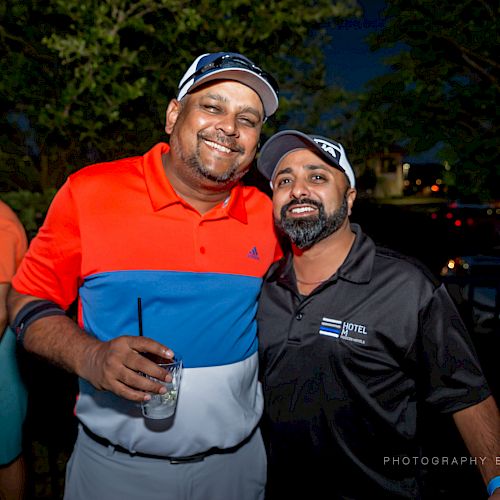 Two people are smiling and posing for a photograph at an outdoor event. One is holding a drink.