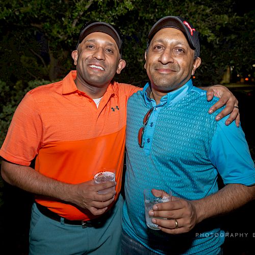 Two men in casual attire, wearing caps and holding drinks, standing close together and smiling for the camera, outdoors at night.