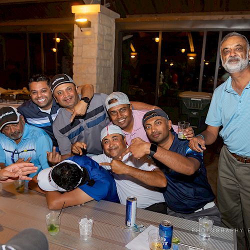 A group of men is posing for a photo at a social gathering, some wearing sports caps and one person is lying on the table.