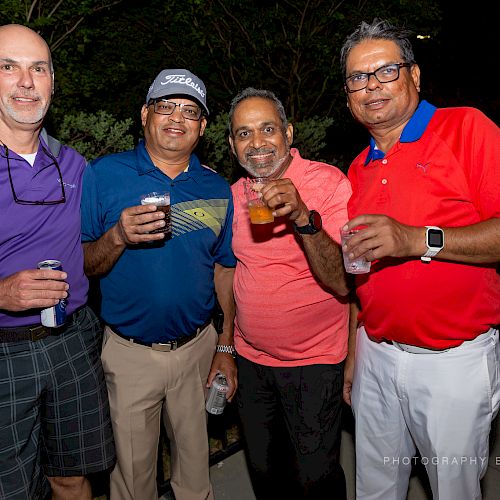 Four men are standing together outdoors, smiling and holding drinks. They are dressed casually and look like they are enjoying a social event.