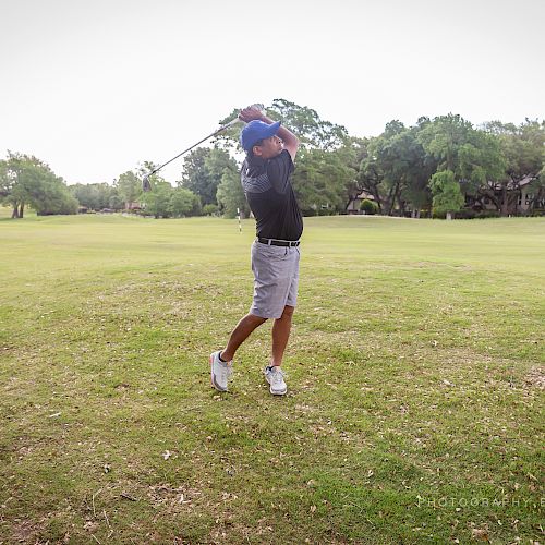 A person is swinging a golf club on a large, well-maintained golf course with trees in the background, wearing shorts and a cap.