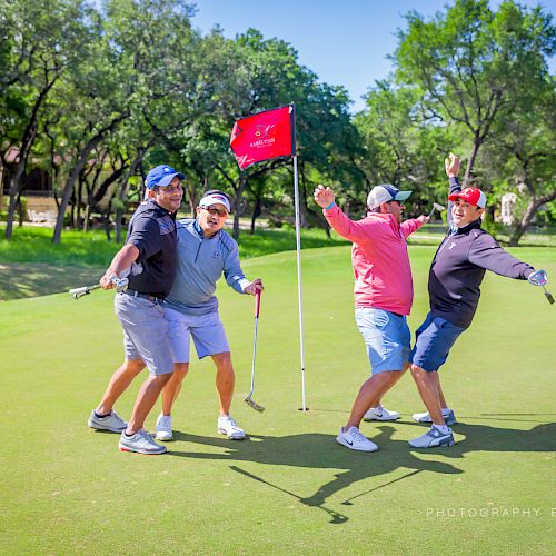 Four people are joyfully posing and celebrating with golf clubs on a golf course near a red flag amidst lush greenery under a clear blue sky.