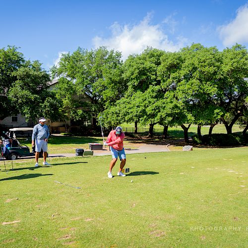 Four people are on a golf course, one is swinging a club while the others watch. It's a sunny day with green trees in the background.