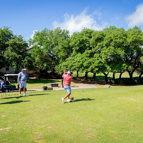 A group of people on a golf course, with one person swinging a golf club while three others watch nearby, surrounded by trees and golf carts.