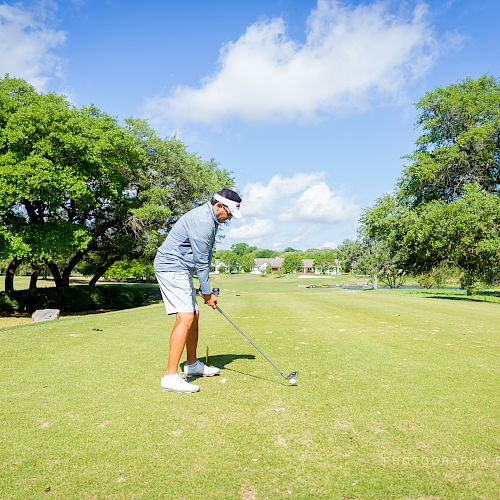 A person is preparing to hit a golf ball on a lush green golf course under a clear blue sky with trees in the background, sunny day.