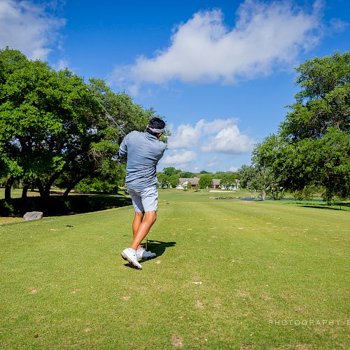 A golfer is mid-swing on a sunny day, surrounded by a green golf course with trees and a clear, blue sky. Ending the sentence.