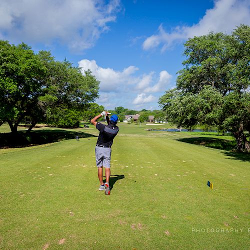 A person is golfing on a sunny day, swinging a club on a lush green course surrounded by trees and blue skies.