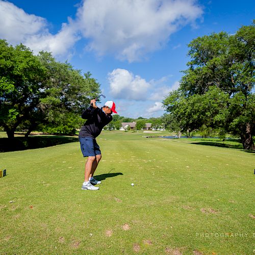 A person is playing golf, swinging a golf club on a green course with trees and a blue sky in the background.