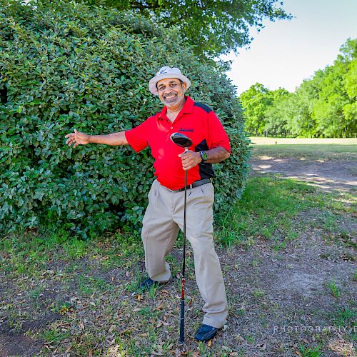 A man in a red shirt and white hat stands outdoors with a golf club, smiling and gesturing toward a bush, on a sunny day.