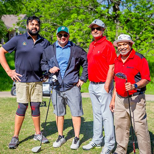 Four men are standing on a golf course, each holding a golf club and smiling at the camera.