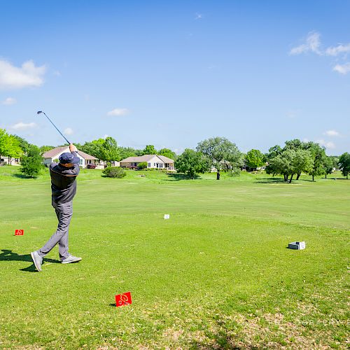 A golfer is teeing off on a lush, green course with trees and houses in the background, under a clear blue sky with a few scattered clouds.