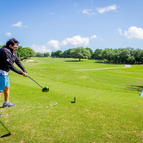 A person is playing golf on a sunny green golf course, about to swing. There's a sign on the right side of the tee box ending the sentence.