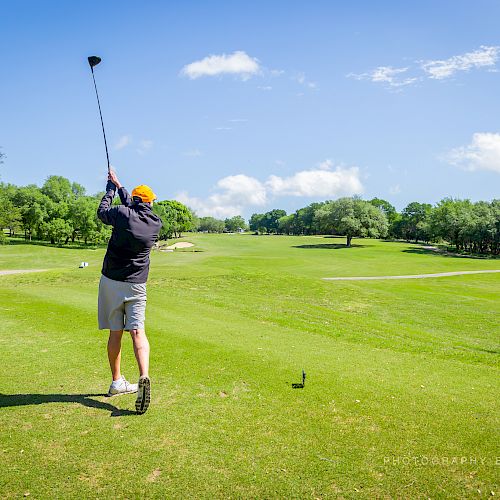 A person is swinging a golf club on a green golf course under a bright blue sky with a few clouds. There is a sign visible on the right.