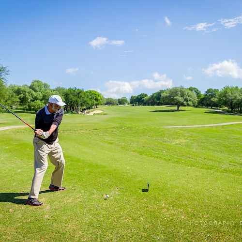 A person is swinging a golf club on a sunny day, standing on a lush green golf course with trees in the background and a sign nearby.