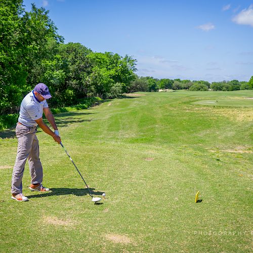 A person is golfing on a lush green course, preparing to swing a club. The sky is clear with a few clouds.