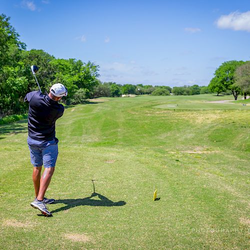 A person is golfing on a lush, green course with trees and clear skies, seen mid-swing with a golf club.