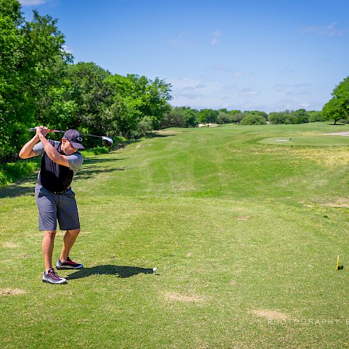 A person is captured mid-swing, playing golf on a sunny day on a well-maintained course with lush greenery and a clear blue sky.