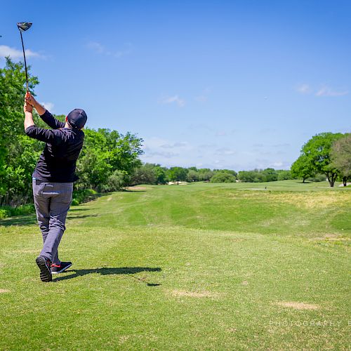 A person in a dark outfit is playing golf, swinging a golf club on a green golf course under a clear blue sky.