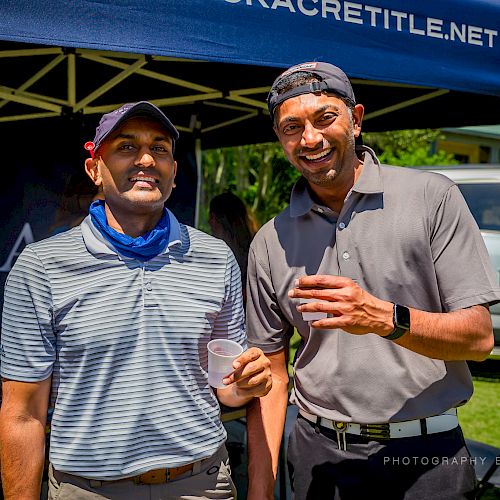 Two people are standing under a canopy, smiling and holding drinks at an outdoor event.