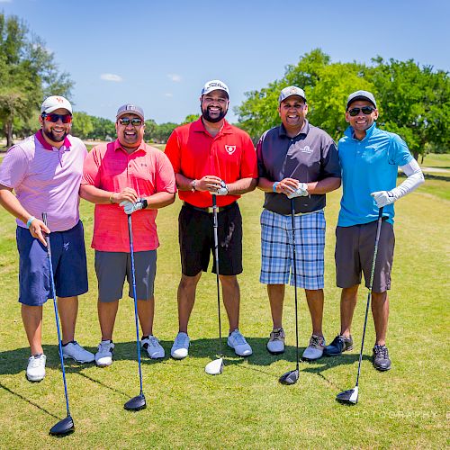 Five men standing on a golf course holding golf clubs, dressed in colorful golf attire under a sunny sky with green trees in the background.