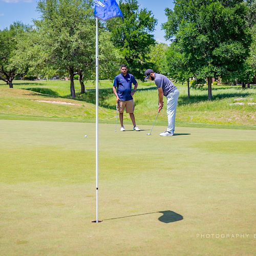 Two people are golfing on a green field, with one putting and the other watching, surrounded by trees and a blue sky above.