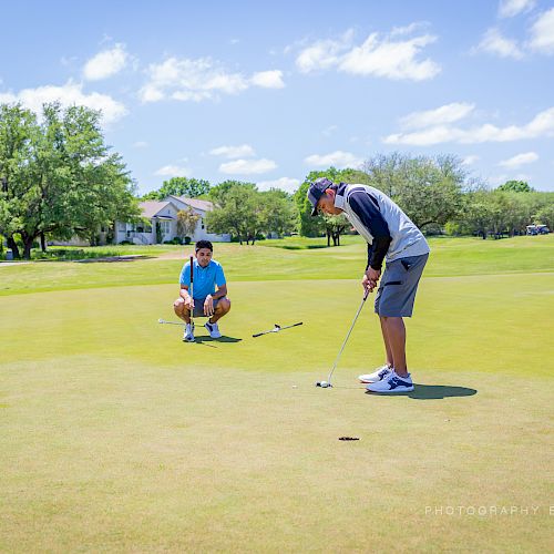 Two people are playing golf on a sunny day; one is lining up a putt while the other kneels behind, observing.