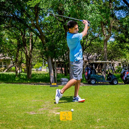 A person wearing a blue shirt swings a golf club on a lush green course with trees and golf carts in the background, ending the sentence.