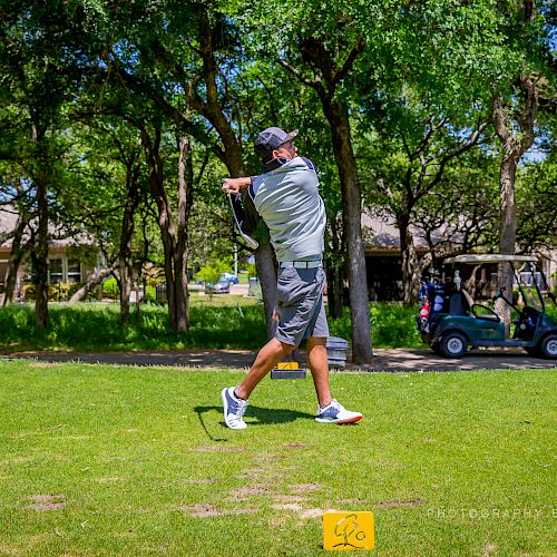 A man is golfing on a lush green course, mid-swing with a golf club, surrounded by trees and several golf carts in the background.