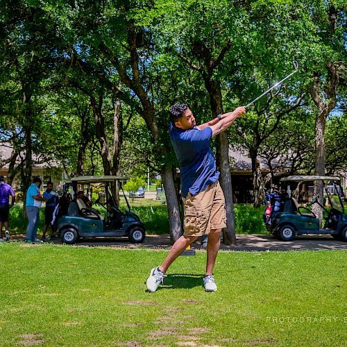 A person is swinging a golf club on a golf course with golf carts and people in the background.