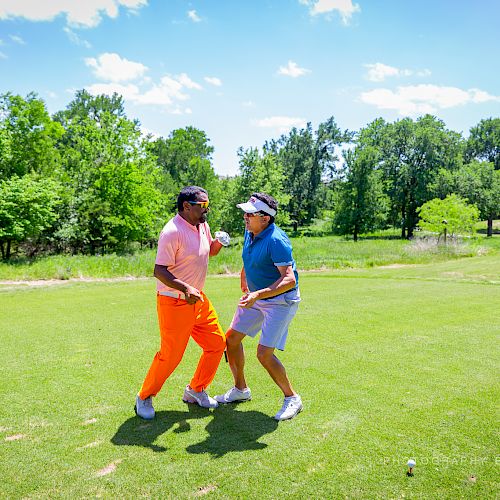 Two individuals in colorful outfits are celebrating on a golf course, surrounded by greenery and a clear, sunny sky in the background.