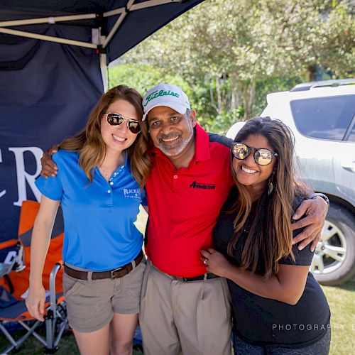 Three people smiling and posing together outdoors in front of a tent and vehicle on a sunny day, with branded items around them.