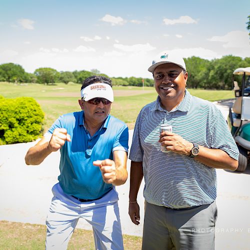 Two men standing on a golf course, one striking a playful pose, and a golf cart in the background under a clear sky.