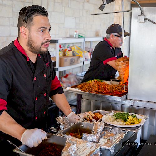 Two chefs are preparing food at a buffet-style setup, serving dishes to customers while wearing black uniforms with red accents.