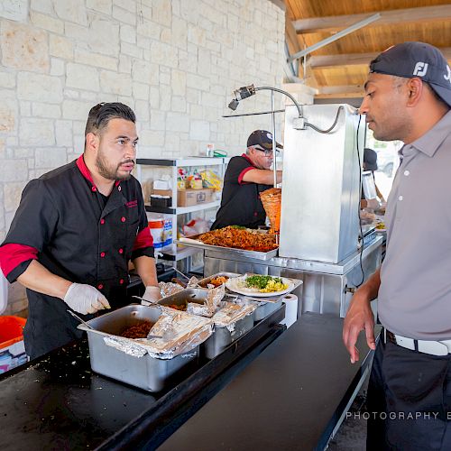 A chef is preparing food at an outdoor counter while a man in a cap and polo shirt watches. A platter of food is on the counter.