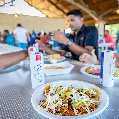 A group of people are gathered at an outdoor event, dining on tacos and drinks at a picnic table under a pavilion.