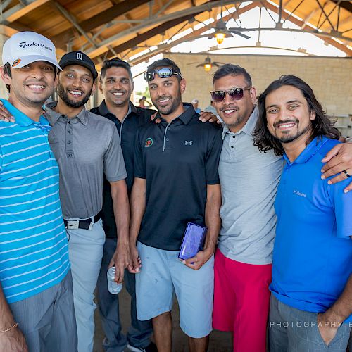 Six men are standing together, posing for a photo in a semi-outdoor setting, likely at an event. They are casually dressed and smiling at the camera.