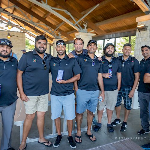 A group of eight men wearing black polo shirts and shorts stand together under a pavilion, some holding drinks, and one holding an award.