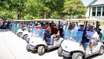 A row of people sitting in golf carts lined up on a sunny day, ready for a golf outing, with trees and a building in the background.