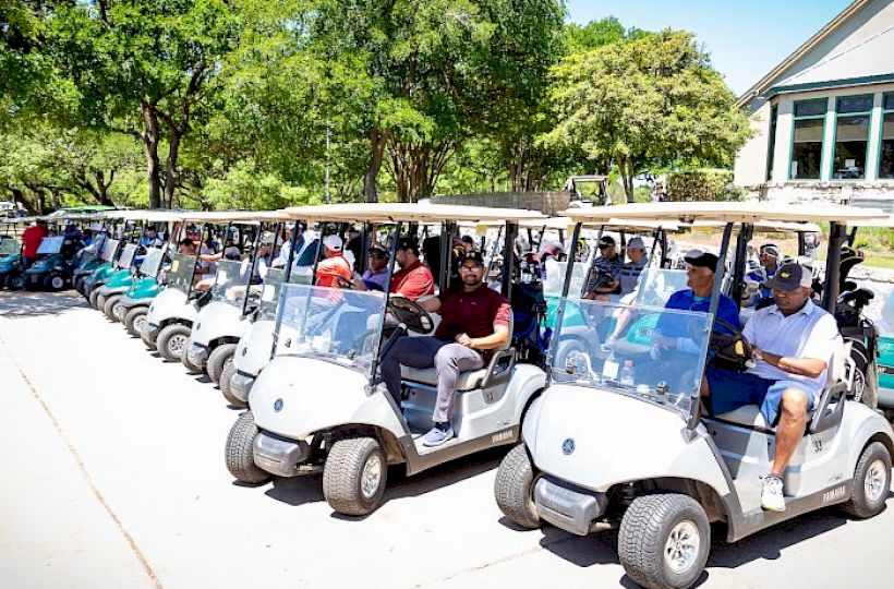 A row of people sitting in golf carts lined up on a sunny day, ready for a golf outing, with trees and a building in the background.