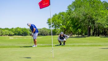 Two people are playing golf on a green course, with one putting and the other squatting and observing near a red flag.