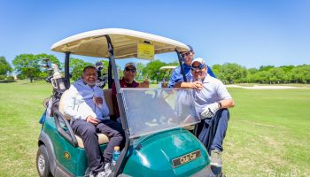 A group of four people are posing cheerfully beside and inside a golf cart on a sunny day at a golf course.