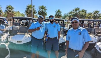 Three men wearing blue shirts and caps are standing in front of golf carts with palm trees in the background, smiling at the camera.