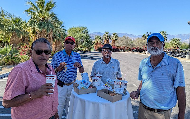 Four men are standing around a table outdoors, holding cups and food boxes, with a scenic background of palm trees and mountains.