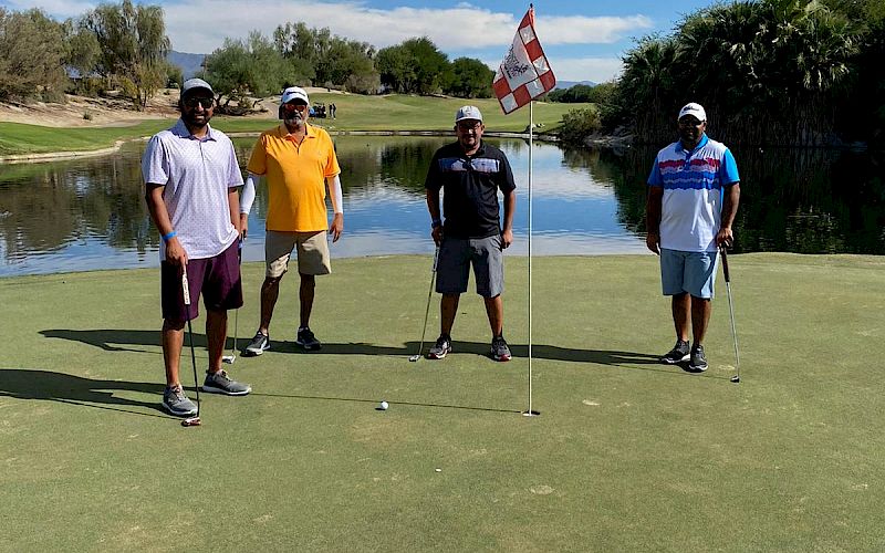 Four people are standing on a golf course green near a hole with a red and white flag, with a scenic lake and trees in the background.