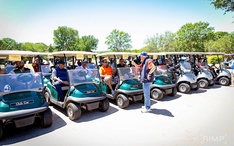 A group of people ready to golf, seated in a line of golf carts, outdoors on a sunny day, with trees in the background.